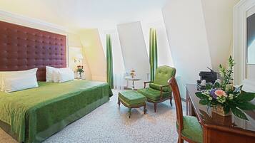 A hotel room with a large bed, a green upholstered chair, a small table with a lamp, and a decorative plant.