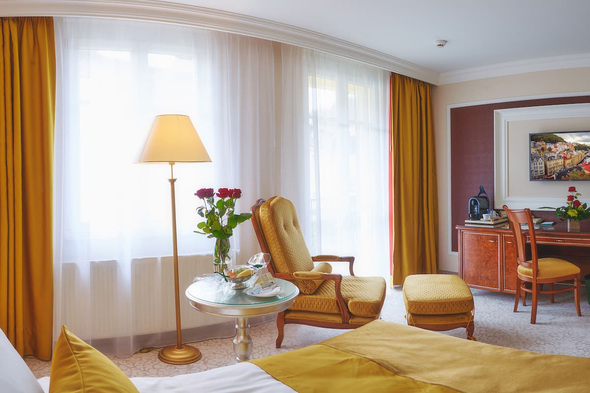 Une chambre d’hôtel comprenant un lit, un coin salon avec un fauteuil et un pouf, un bureau avec une chaise, une lampe et un tableau au mur.