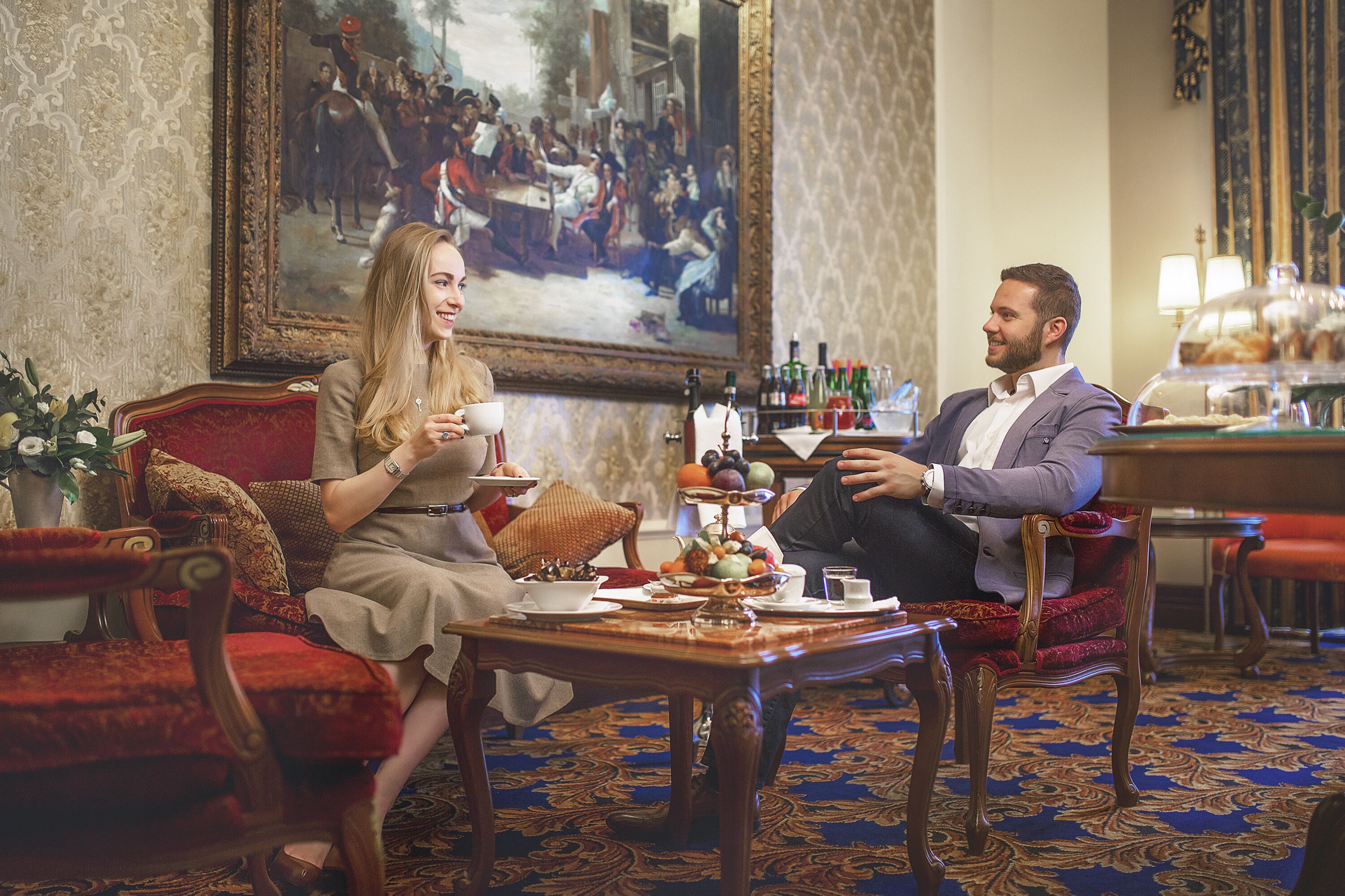 A room with a large painting, a dining table with a fruit bowl, and a red upholstered chair.