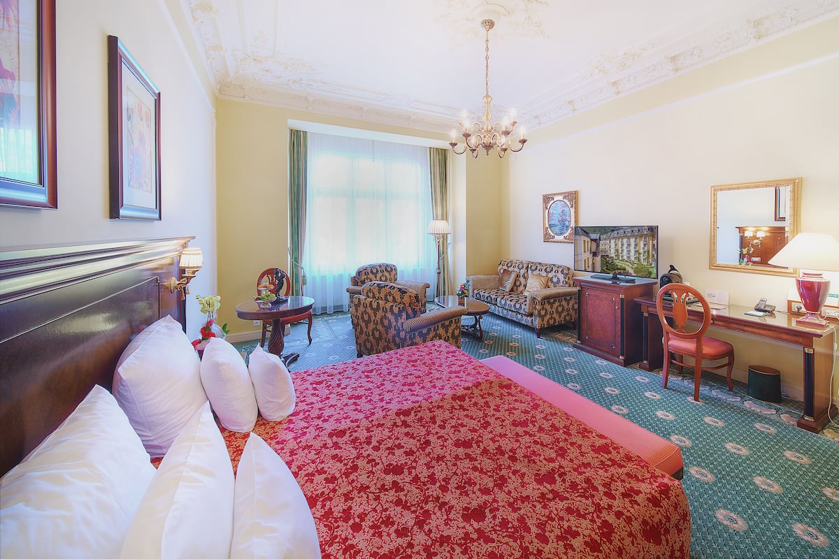 Une chambre d’hôtel avec un grand lit, un canapé, un bureau et une chaise. On y trouve un lustre, une fenêtre avec des rideaux et un tableau au mur.