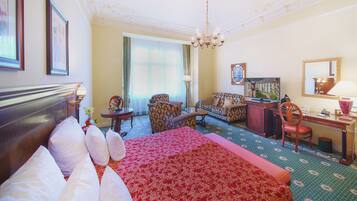 Habitación de hotel con una cama grande, un sofá, un escritorio y una silla. Hay una araña, una ventana con cortinas y un cuadro en la pared.