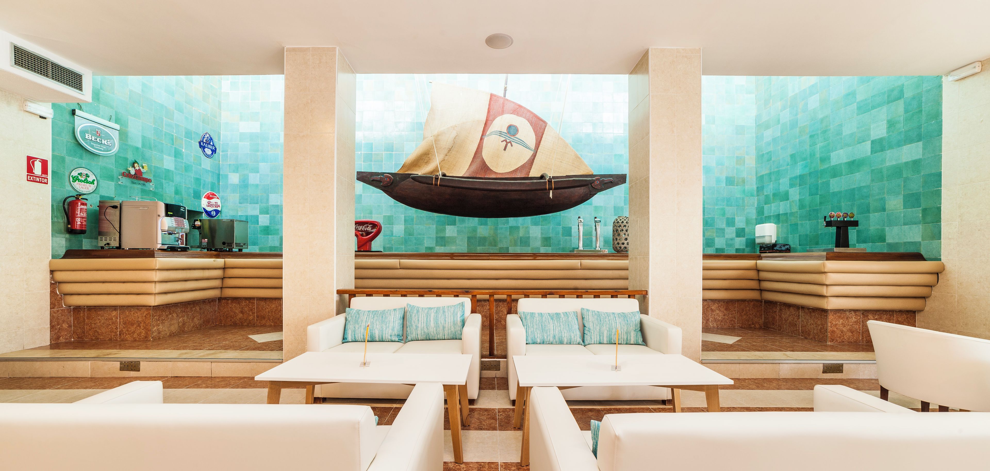 Een moderne hotellobby met een centraal zitgedeelte, een hangende bootdecoratie en een betegelde muur met verschillende logo's.