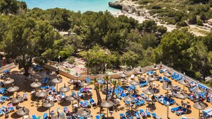 A coastal resort with a beach area featuring sun loungers and umbrellas, a view of the sea, and buildings in the background. - Globales Samoa (Manacor)