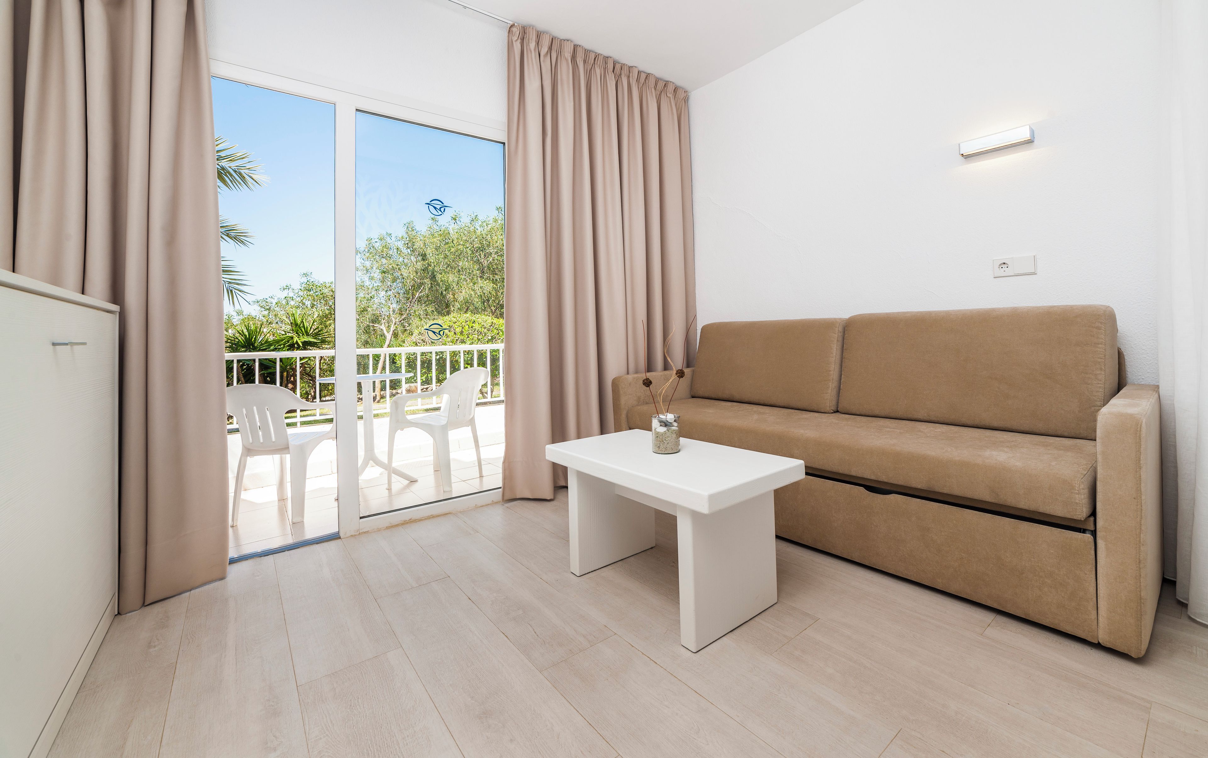 Un soggiorno moderno con un divano beige, un piccolo tavolo bianco e un balcone con sedie bianche.