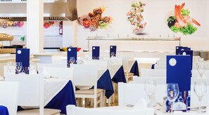 A restaurant with tables set for dining, a kitchen area, and decorative wall art. - Globales Samoa (Manacor)