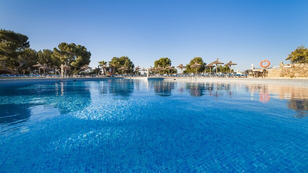 A clear blue pool with lounge chairs and umbrellas, surrounded by trees and a stone wall. - Globales Samoa (Manacor)
