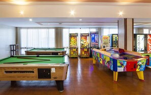 Una mesa de billar, máquinas de videojuegos y una mesa de tenis de mesa colorida en una habitación con suelo de madera y ventanas grandes.