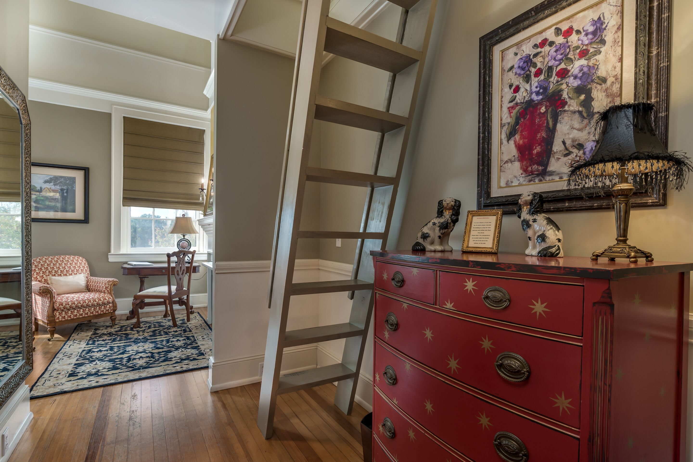 A room with a wooden floor, a red dresser with star-shaped handles, a ladder, a chair, a framed painting, and a lamp.