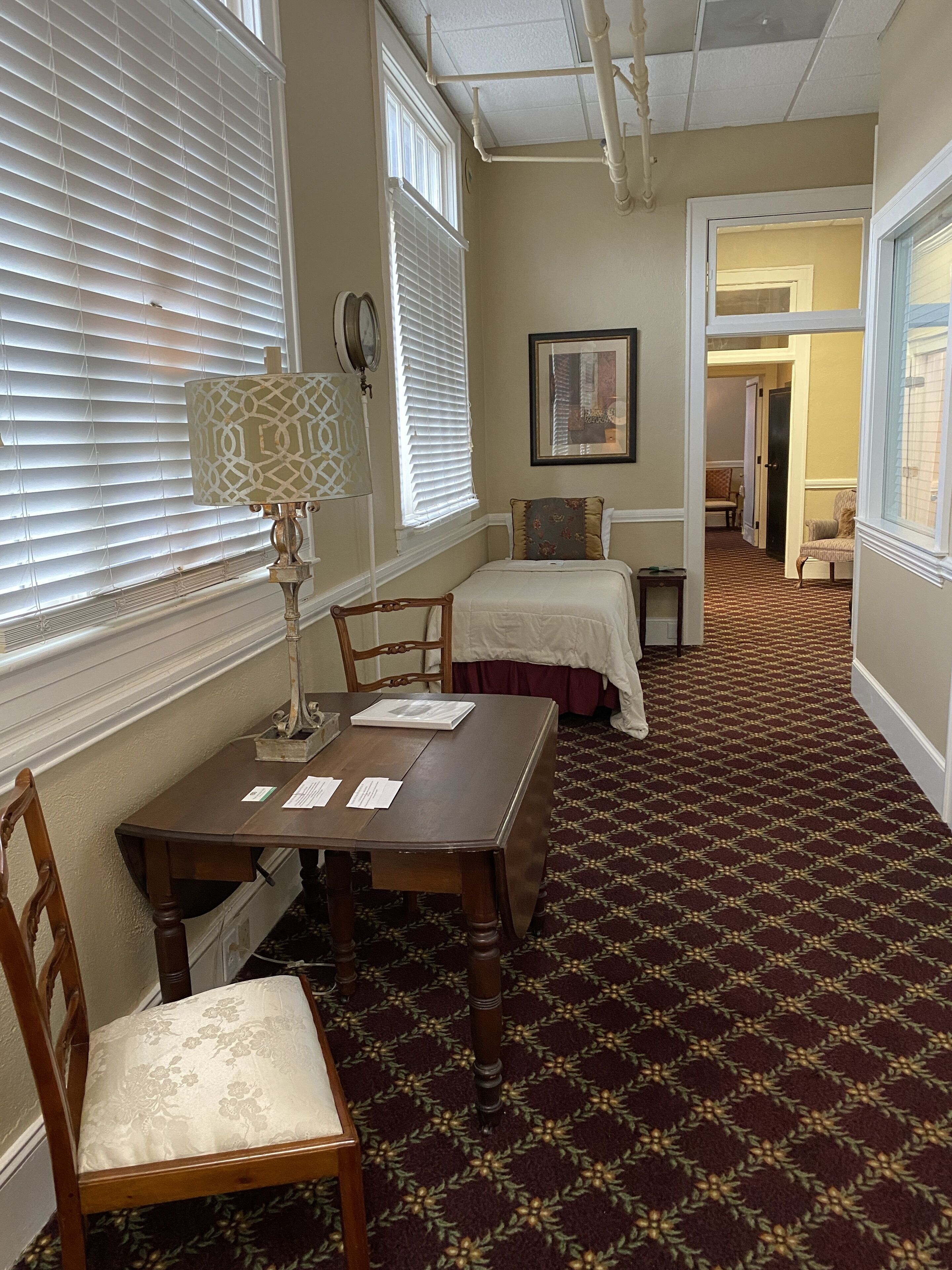A hotel room with a wooden table, two chairs, a lamp, a bed with a white bedspread, and a patterned carpet.