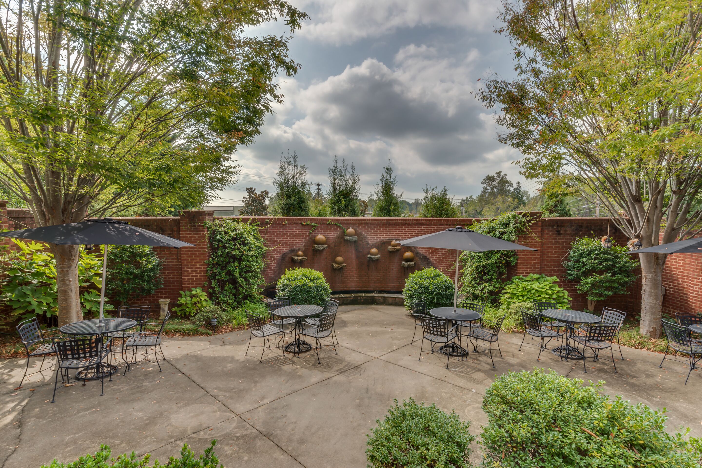 An outdoor patio with round tables and chairs, surrounded by brick walls and greenery.