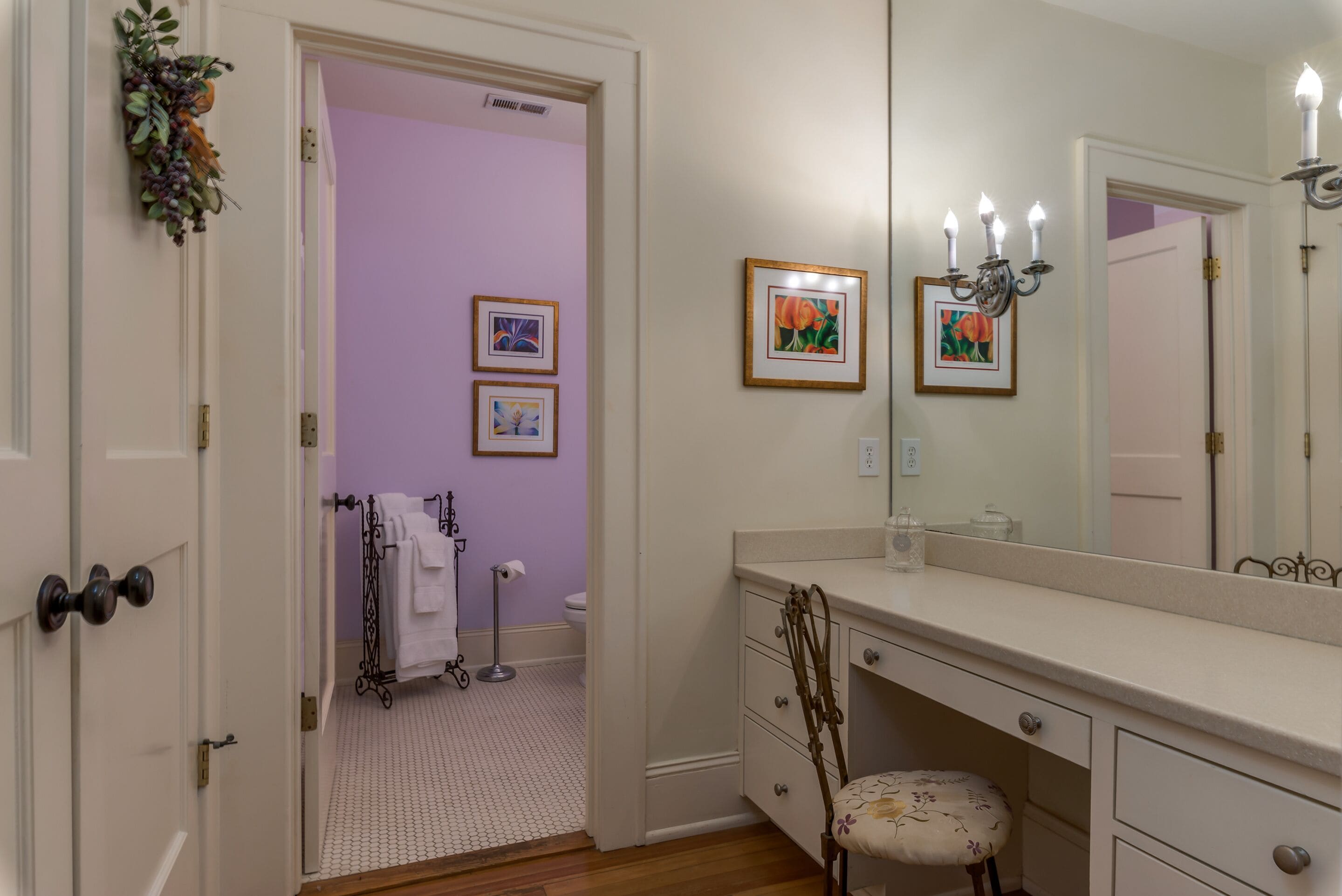 Une salle de bain avec un meuble-lavabo blanc, un grand miroir et des toilettes. Des tableaux encadrés ornent le mur et une plante décorative est posée sur la porte.