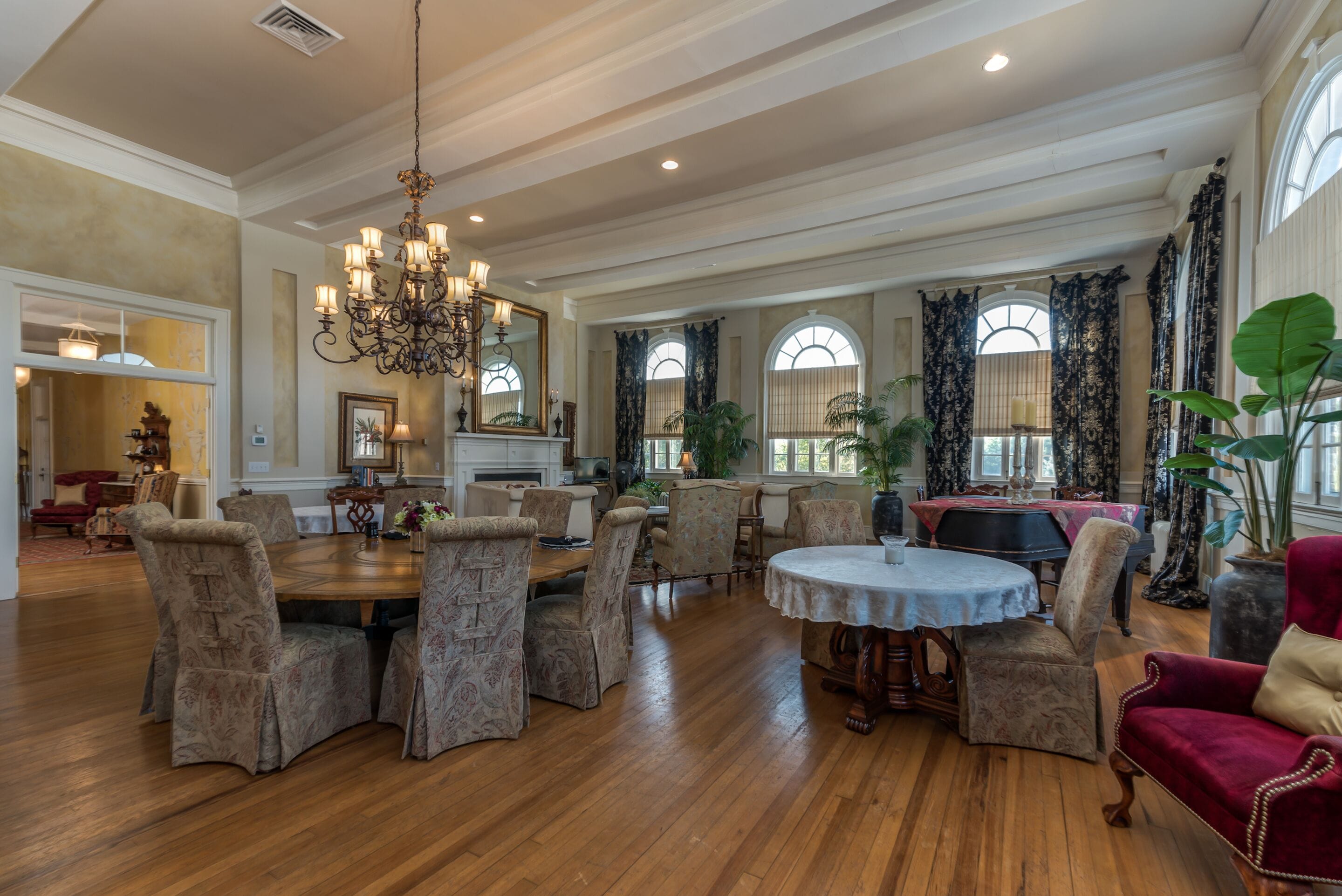 A spacious dining area with a large wooden table surrounded by upholstered chairs, a chandelier, and large windows with curtains.