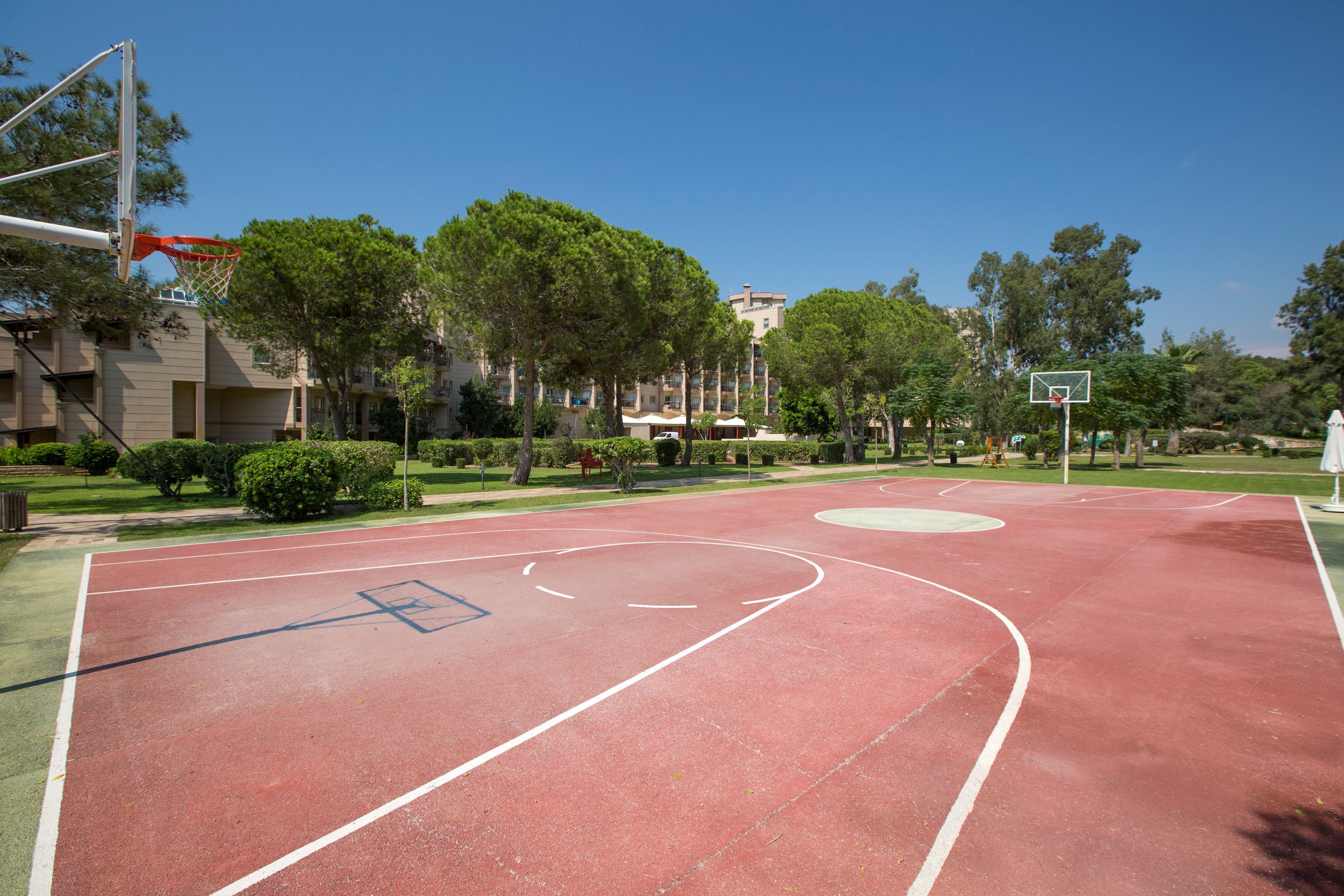 Una cancha de baloncesto al aire libre con superficie roja, rodeada de árboles y un edificio al fondo.