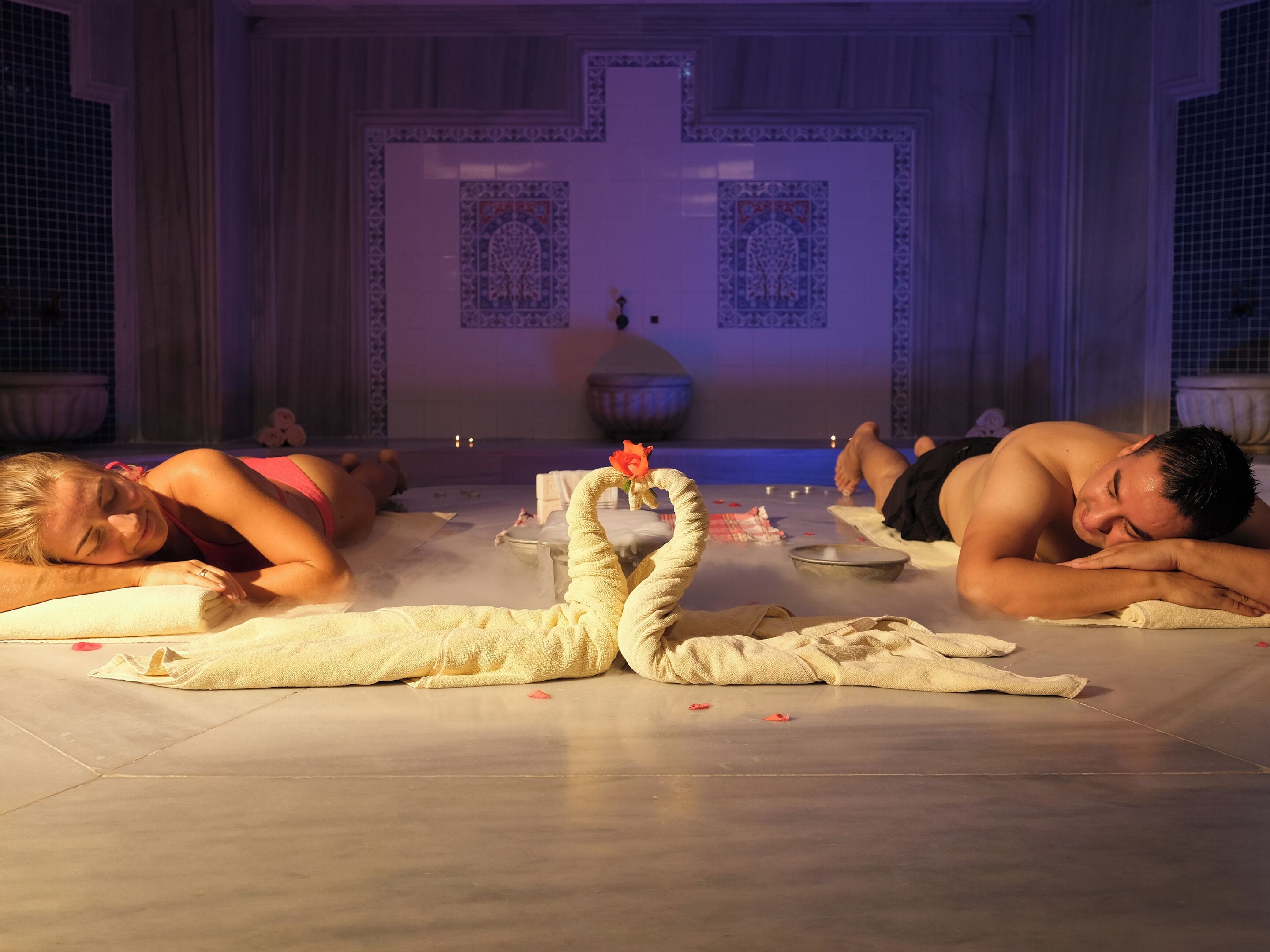 Un cuarto de spa con dos personas acostadas sobre esterillas, una serpiente decorativa y una pared de azulejos con diseños elaborados.