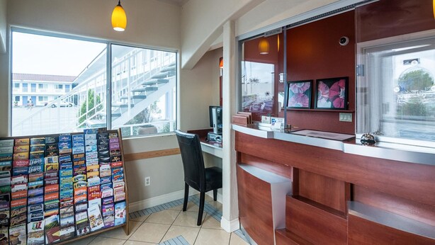 A reception area with a wooden desk, a computer, and a chair. There is a window with a view of a building and a street. - Motel 6 Norwalk, CA (Norwalk)