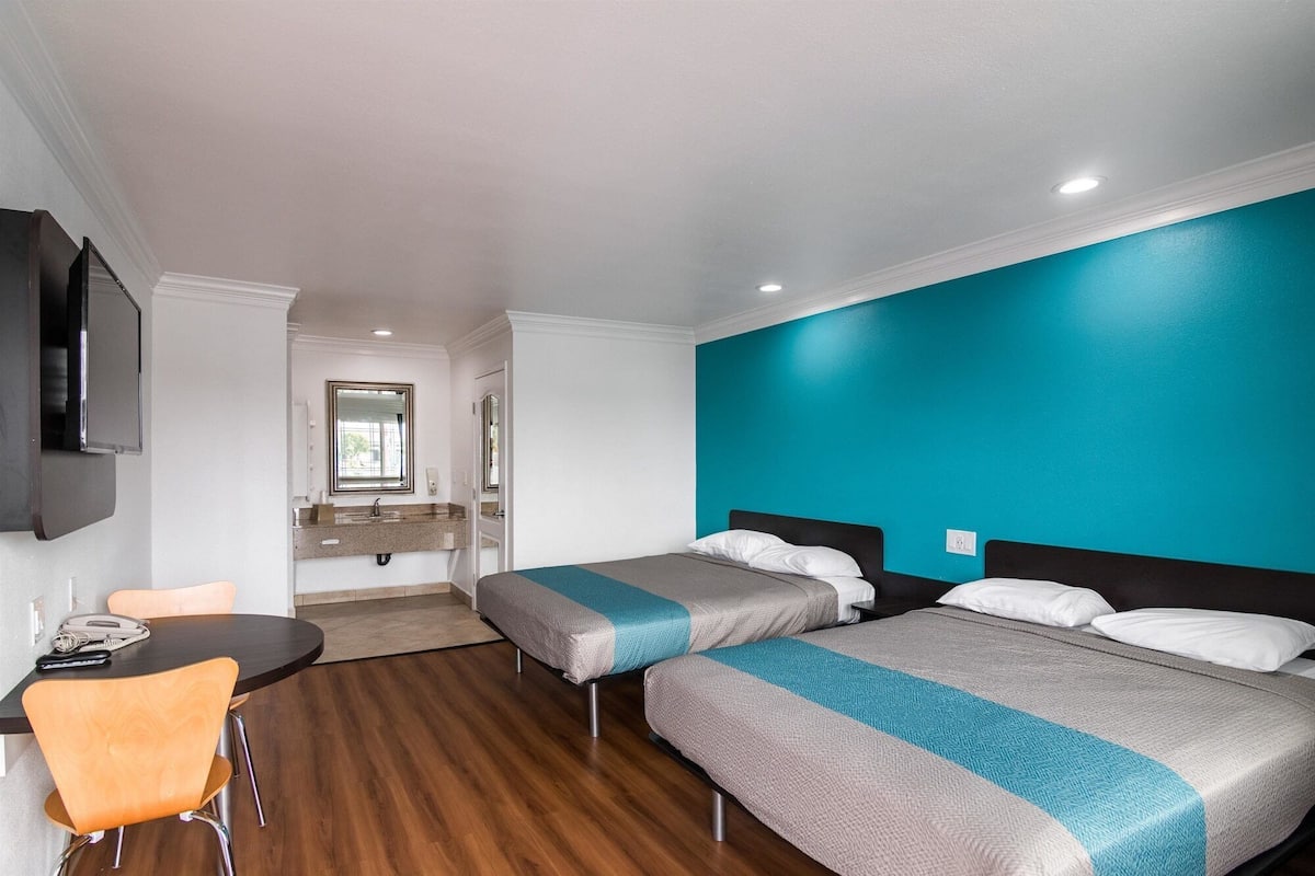 A hotel room with two beds, a desk with a chair, a TV mounted on the wall, and a bathroom visible through an open door.