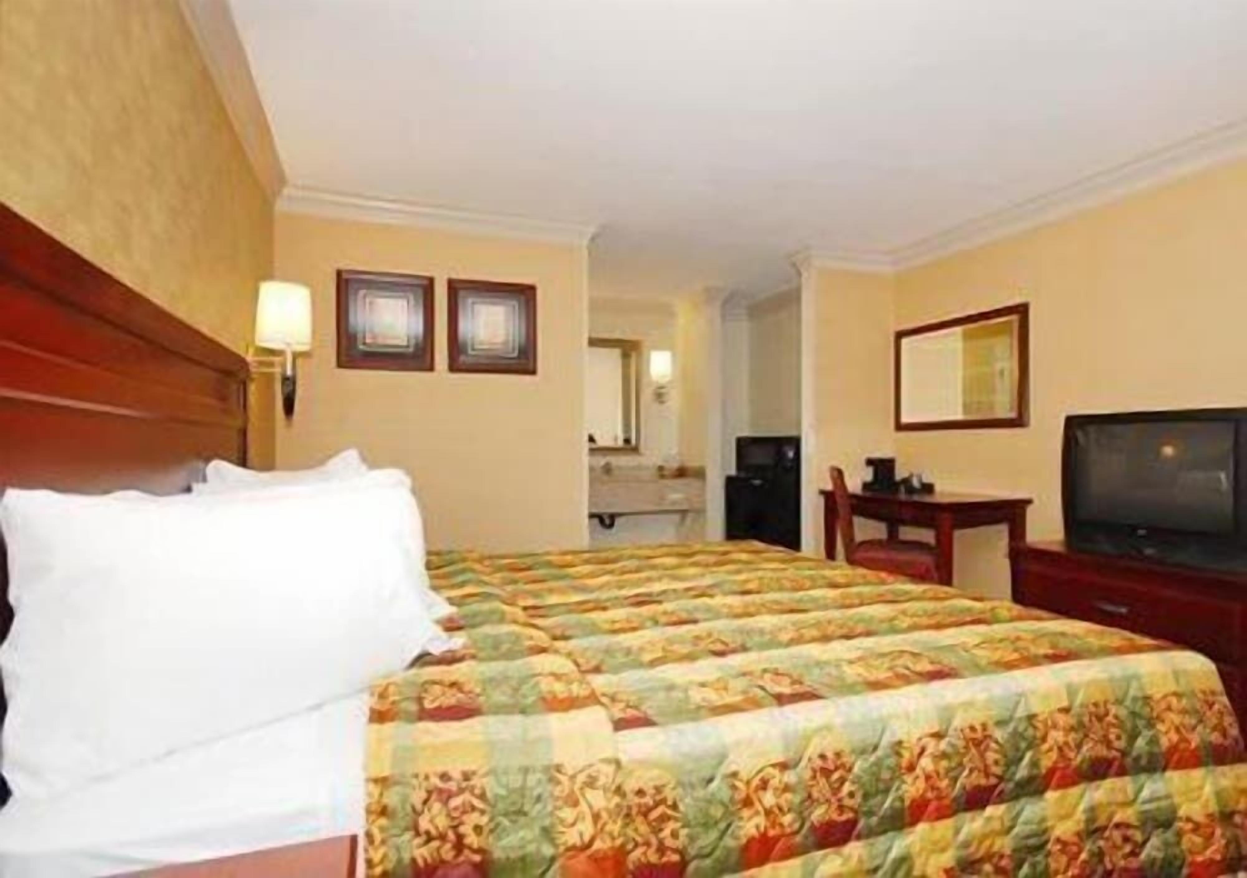 Ein Hotelzimmer mit einem großen Bett, einem Schreibtisch, einem Fernseher und einem im Hintergrund sichtbaren Badezimmer.