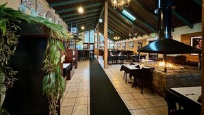 An indoor dining area with a long counter, tables, and chairs, a stone fireplace, and hanging lights.