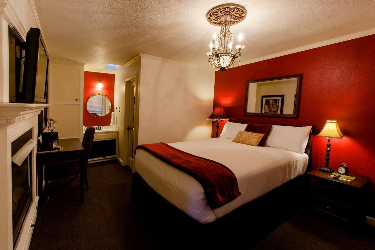 A hotel room with a large bed, a chandelier, a red accent wall, a mirror, and a bedside table with a lamp.