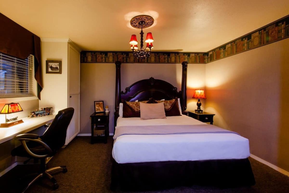 A hotel room with a four-poster bed, a desk with a chair, a desk lamp, a bedside table with a lamp, and a picture on the wall.