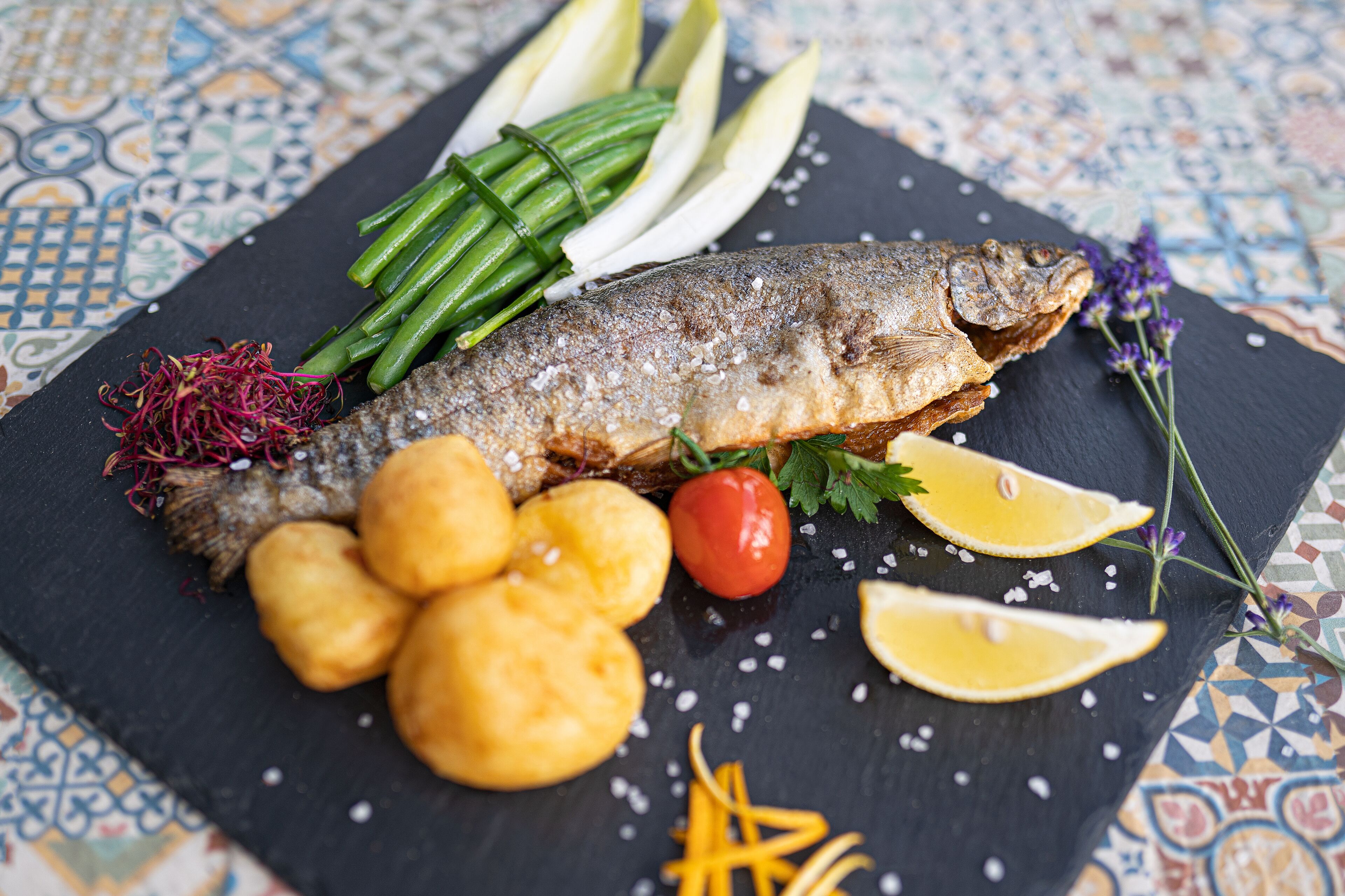 A fish dish with lemon slices, green beans, and a small tomato on a slate plate.