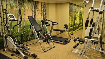 Een fitnessruimte met diverse fitnessapparaten, waaronder een loopband, een crosstrainer en krachttoestellen, met een achtergrond van een bosdecor.