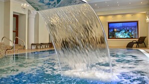 Una amplia piscina interior con una cascada, un área de descanso con sillas y un ventanal con vistas nocturnas.
