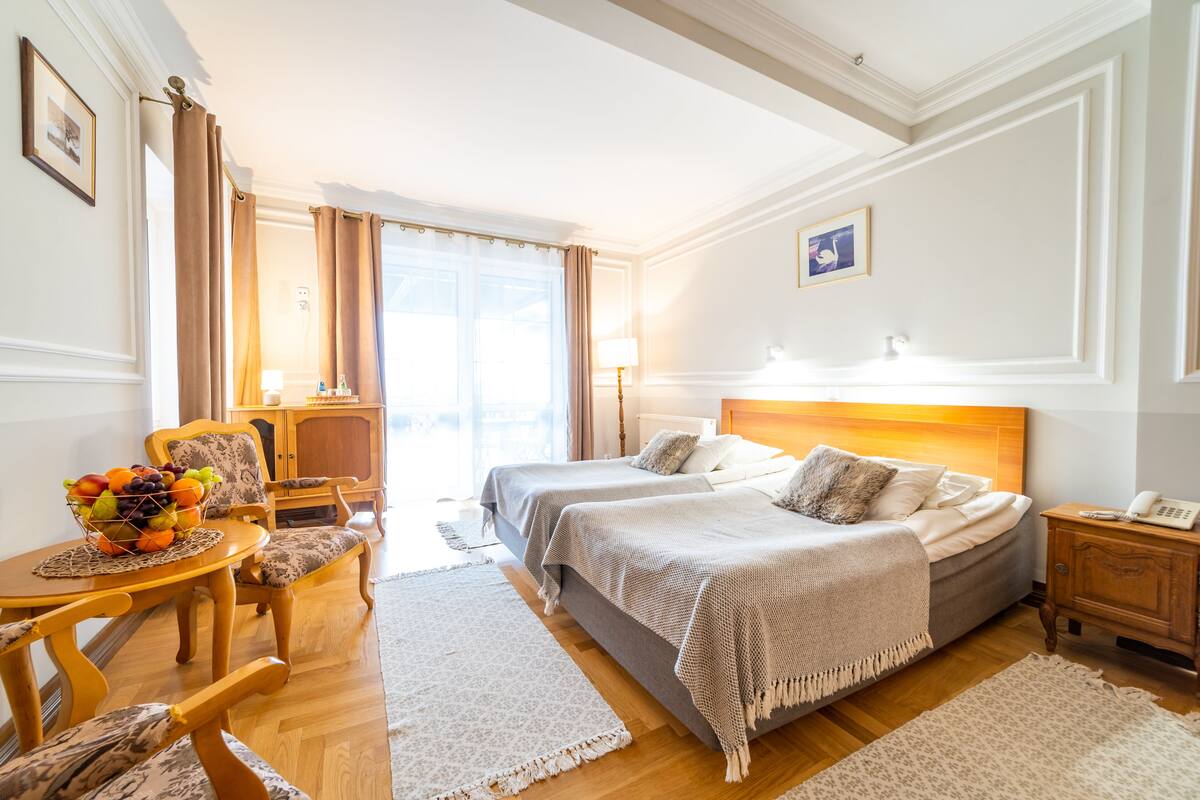 Habitación de hotel con dos camas, un área de comedor con una cesta de fruta, muebles de madera y vistas al exterior.