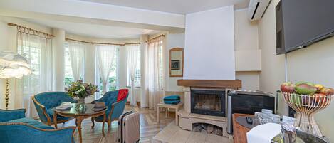 A living room with a fireplace, a flat-screen TV, and a dining area with blue upholstered chairs and a table.