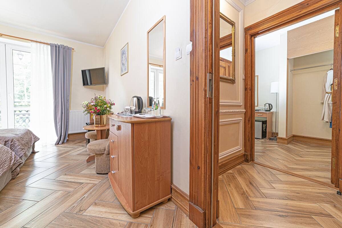 Habitación de hotel con suelo de madera tipo espiga, cómoda de madera, cama, televisor montado en la pared y baño visible a través de una puerta abierta.