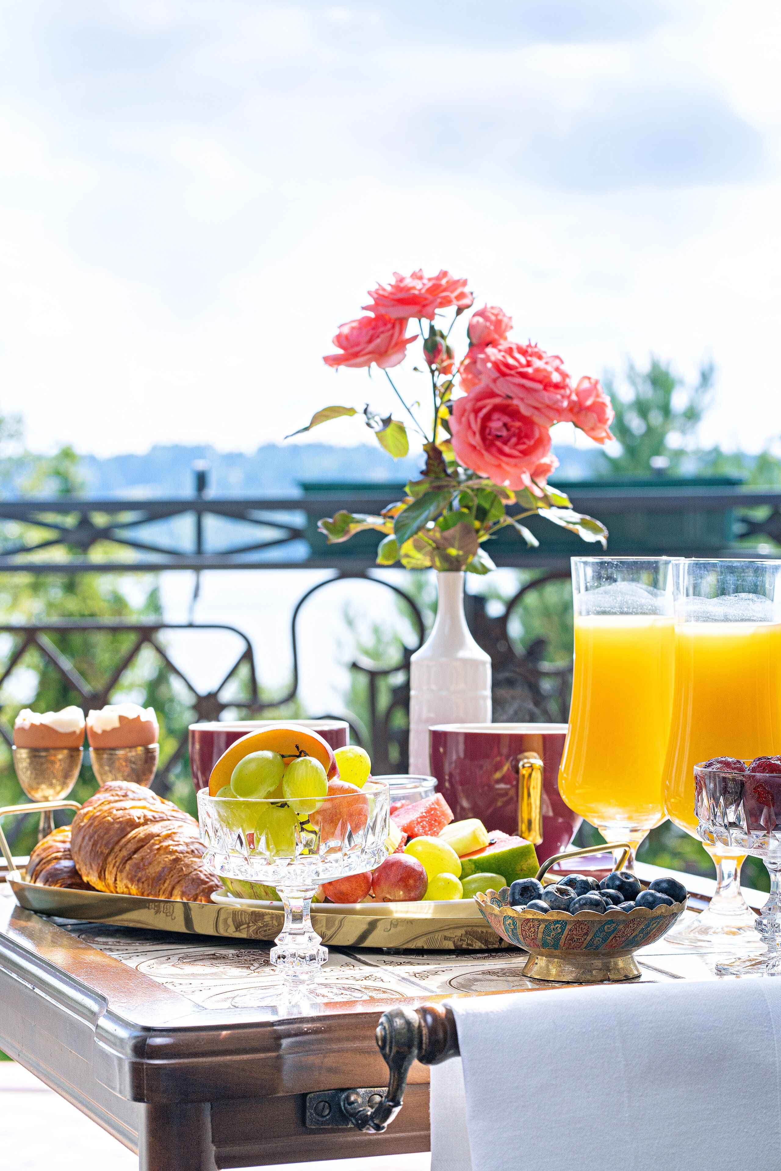 A breakfast table with croissants, fruit, and juice, set outdoors with a view of greenery and a railing.