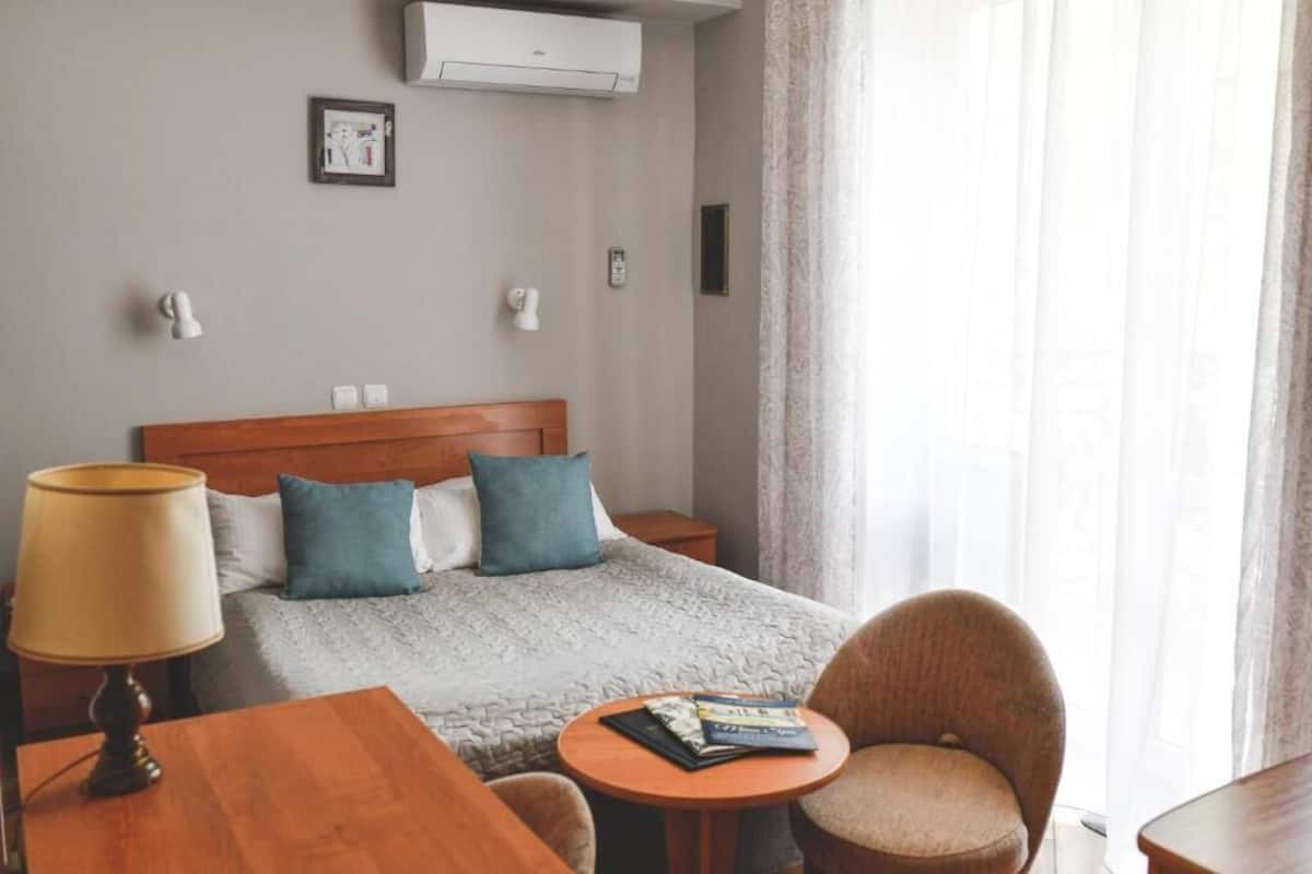 Una habitación de hotel con cama, mesitas de noche, lámpara, sillón, una pequeña mesa y ventana con cortinas.