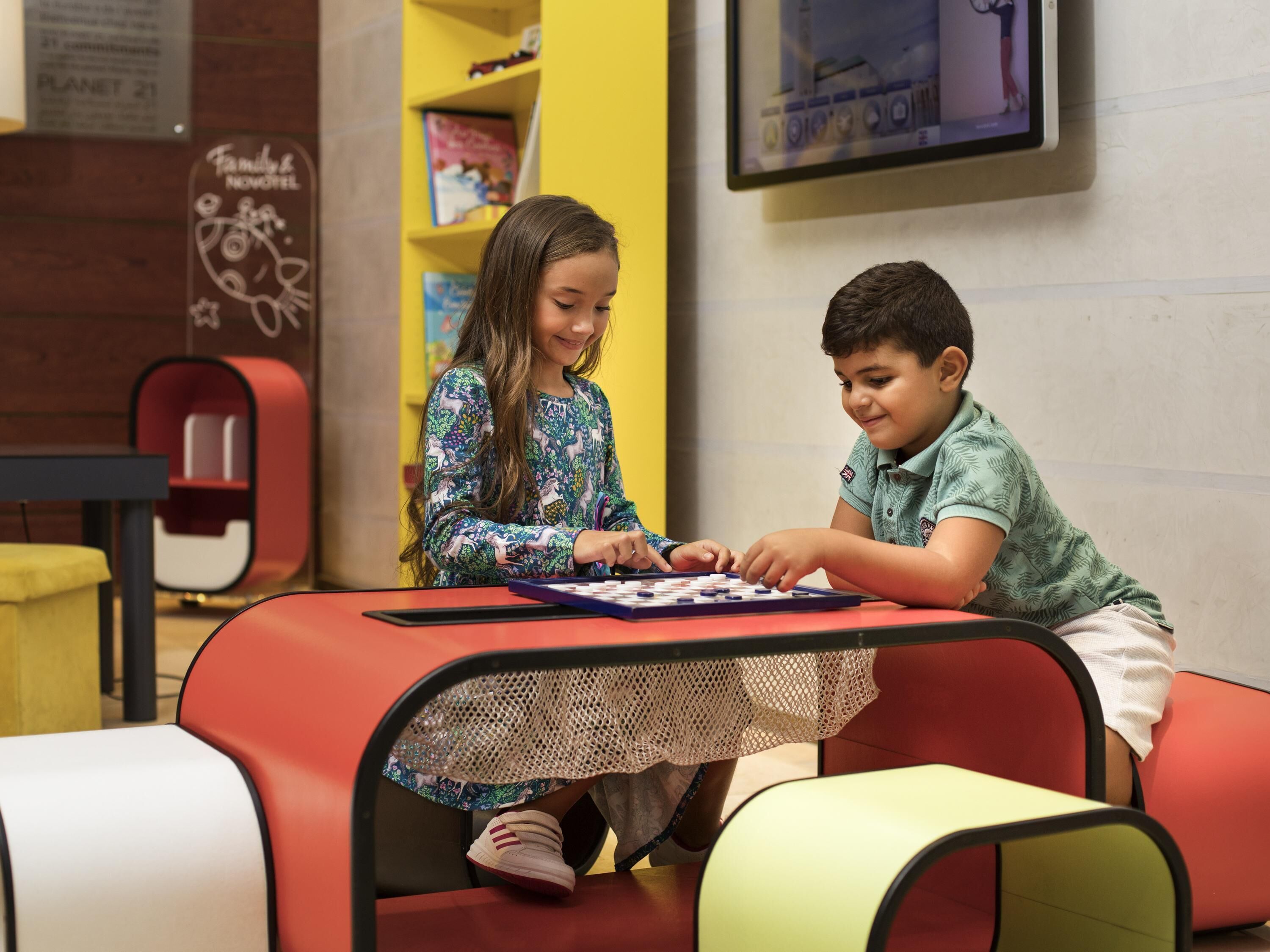 Two children playing a board game at a table in a brightly colored room with bookshelves and a television.