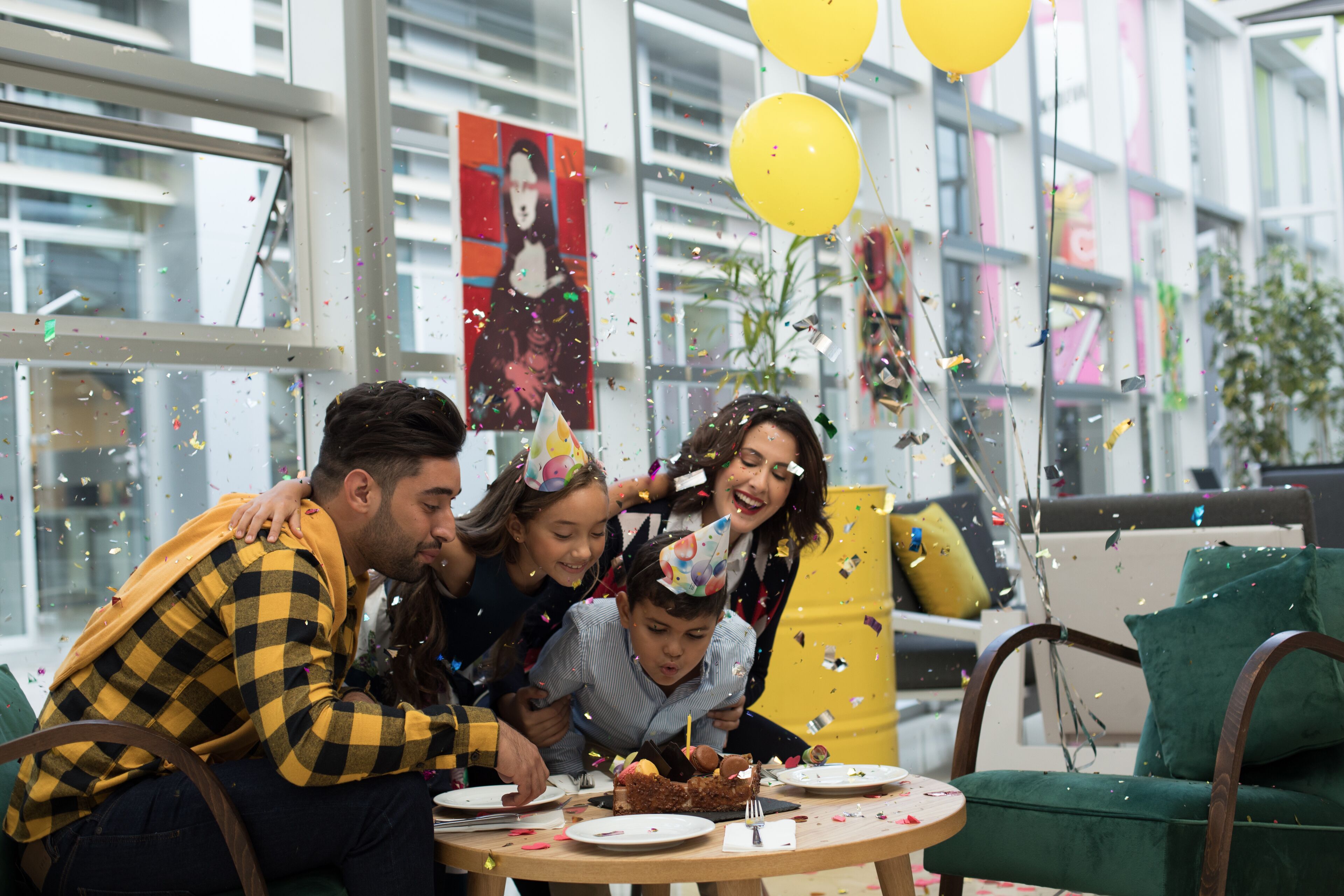 Un grupo de personas celebrando alrededor de un pastel de cumpleaños, con confeti, en una habitación con ventanas grandes y una silla verde.