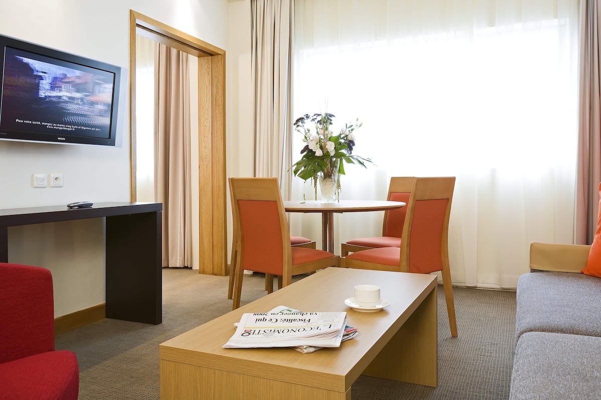 A hotel room with a TV, a dining table with chairs, a sofa, and a window with curtains.