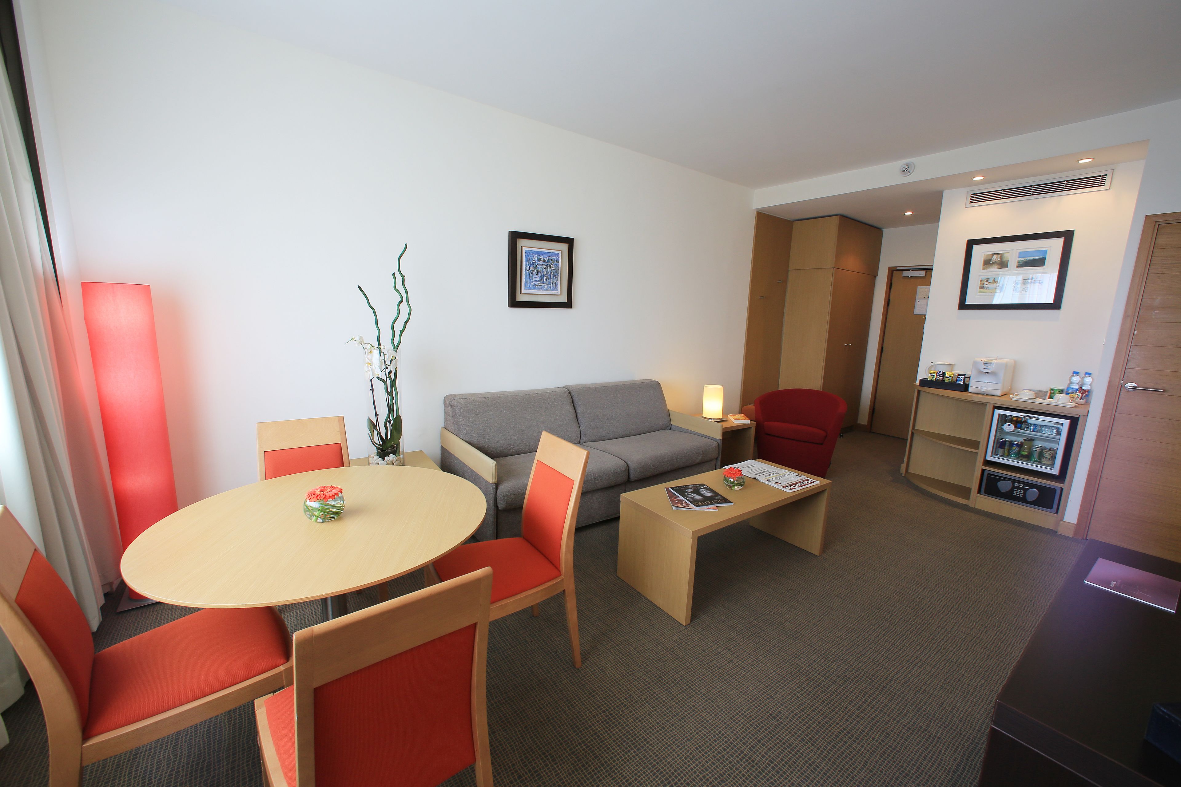 A compact hotel room with a sofa, a dining table with chairs, a small coffee table, a refrigerator, and a TV.