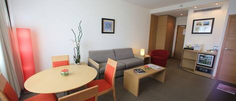 A compact hotel room with a sofa, a dining table with chairs, a small coffee table, a refrigerator, and a TV.