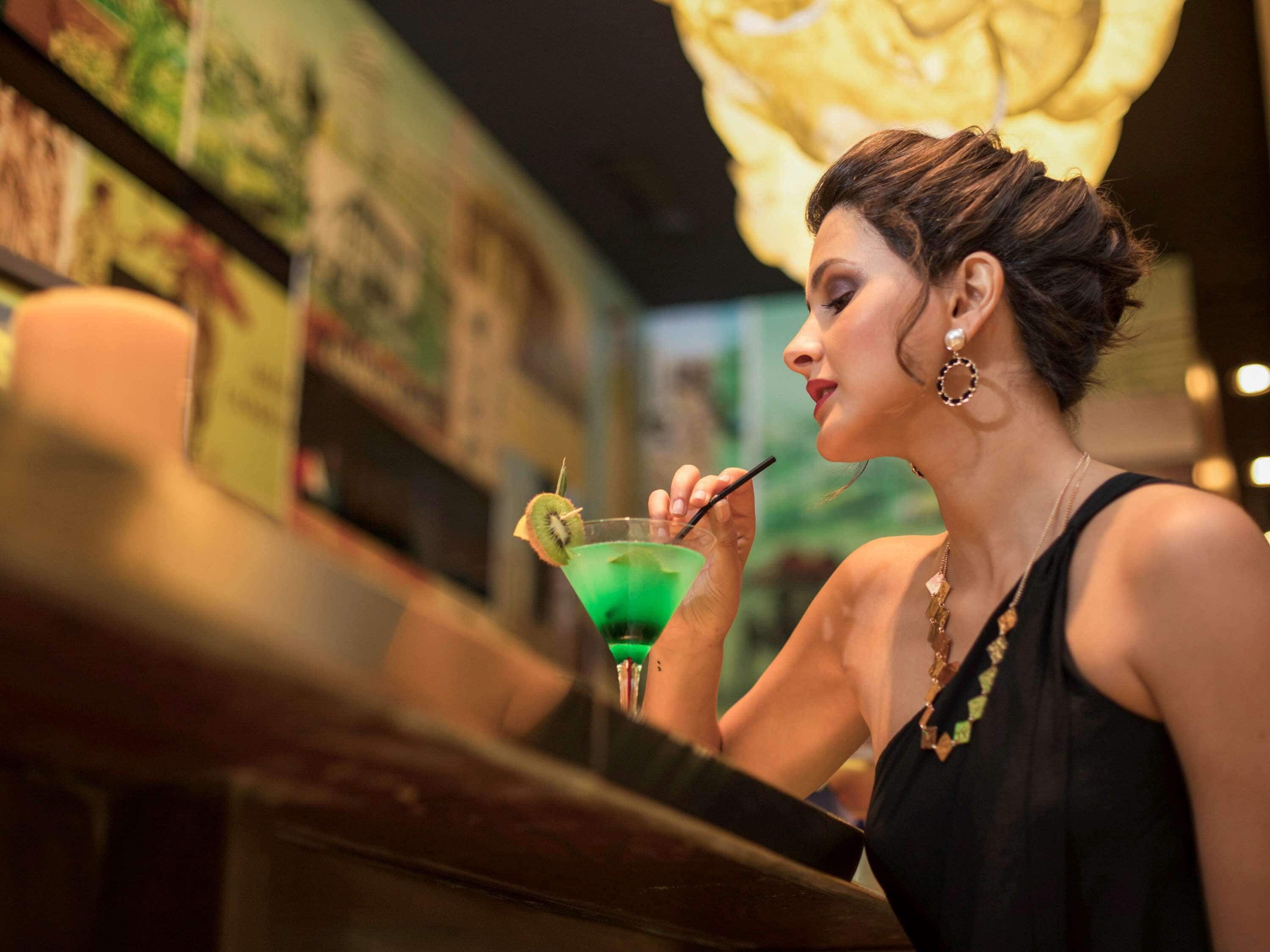 Una persona sentada en la barra con un cóctel verde, un menú y una pared decorada con obras de arte.