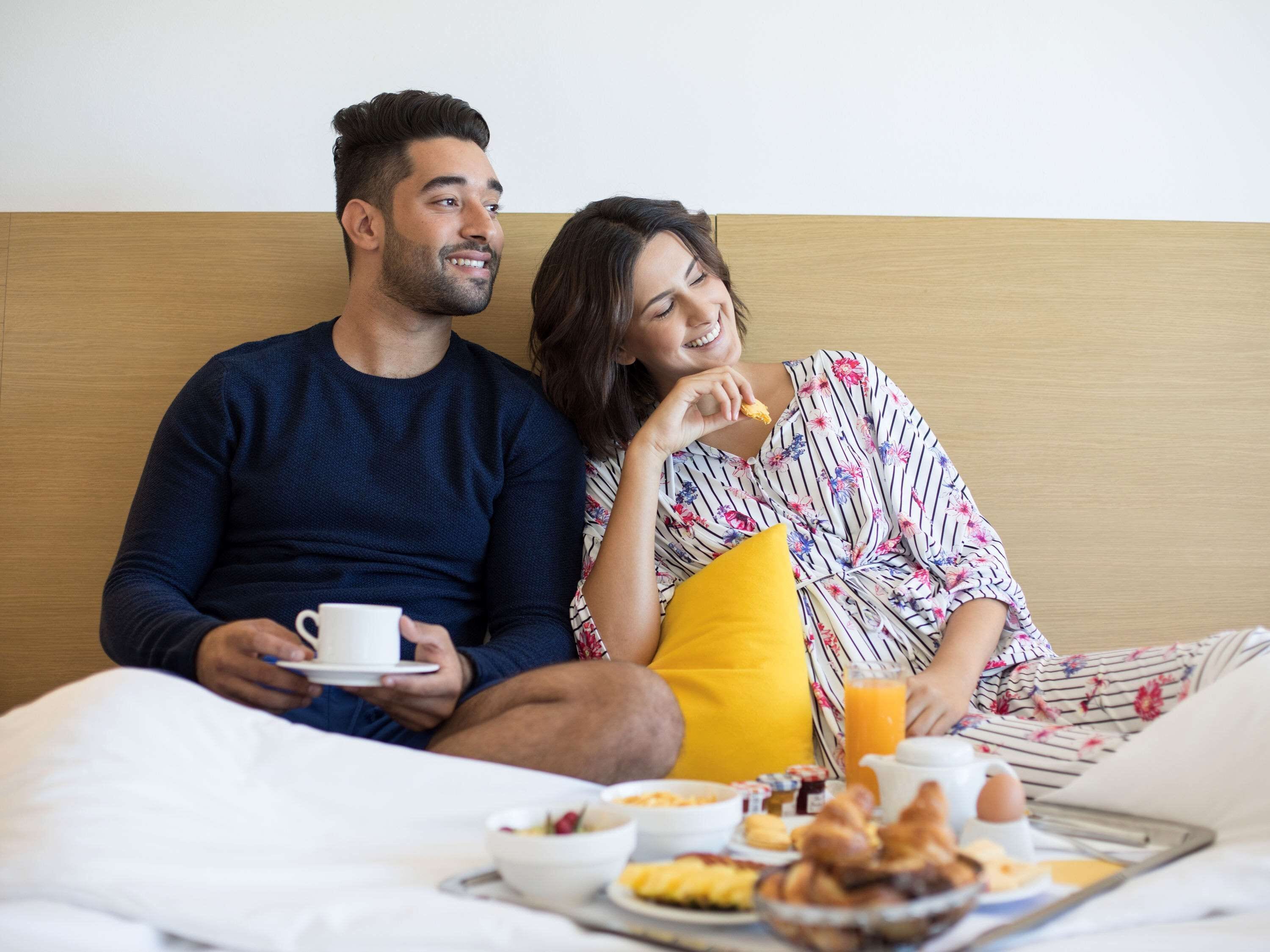 Una pareja sentada en una cama con cosas para el desayuno, incluyendo un tazón de cereal, croissants y jugo de naranja.