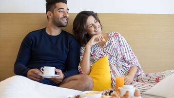 Una pareja sentada en una cama con cosas para el desayuno, incluyendo un tazón de cereal, croissants y jugo de naranja.