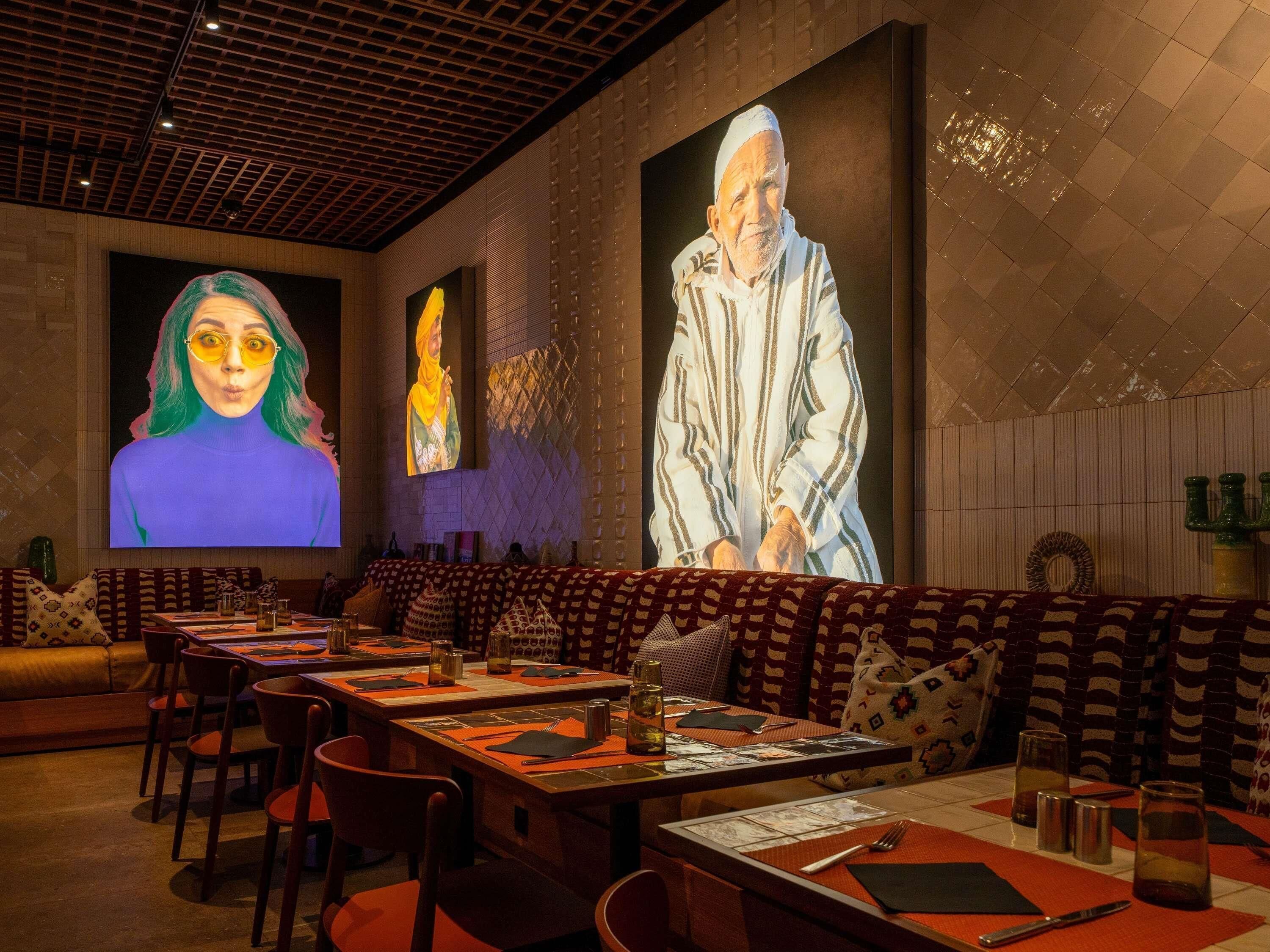 A restaurant with long communal tables, cushioned seating, and large wall-mounted screens displaying images.