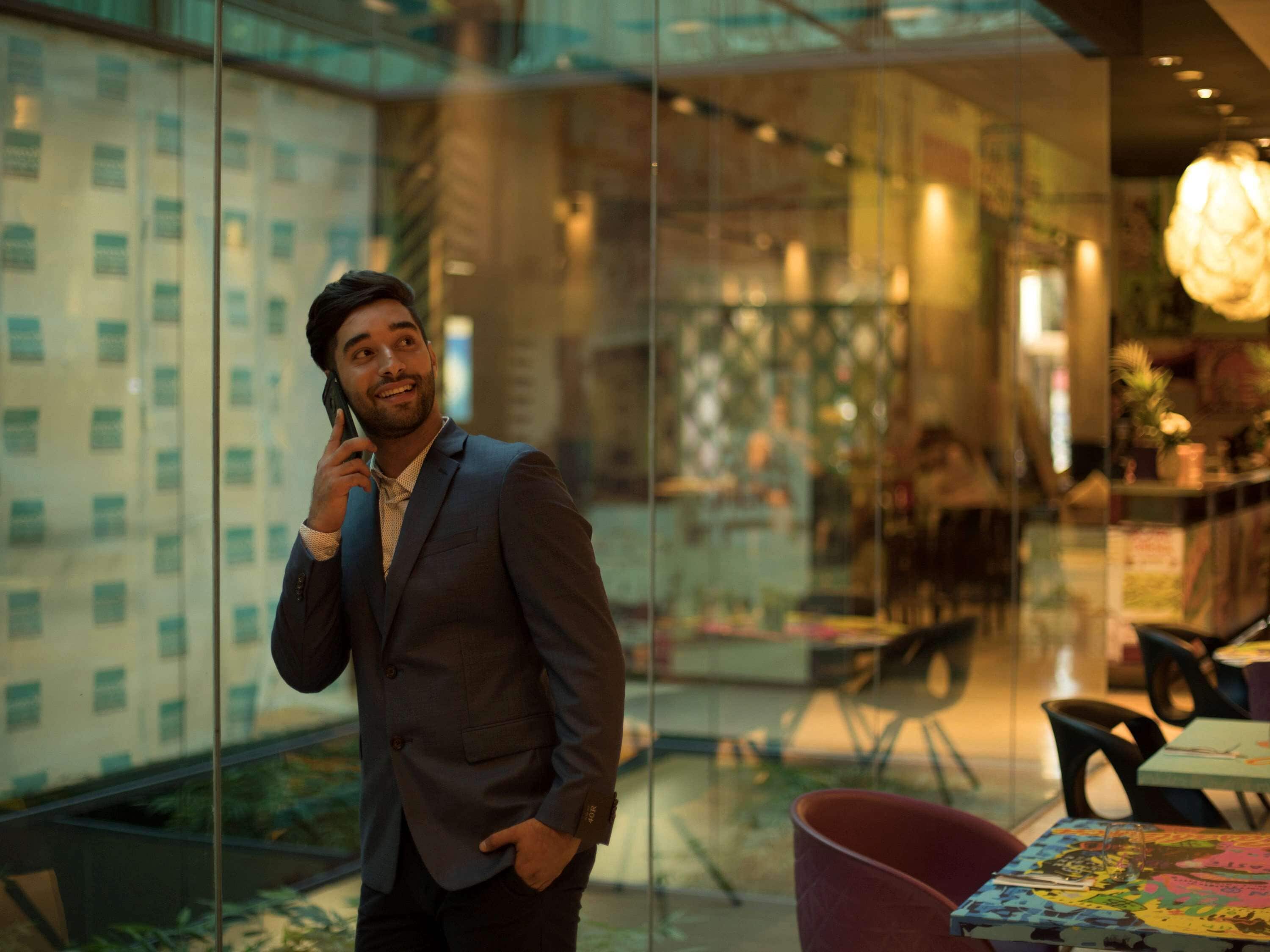 A man in a suit talking on a phone inside a modern building with glass walls, a dining area, and a chandelier.