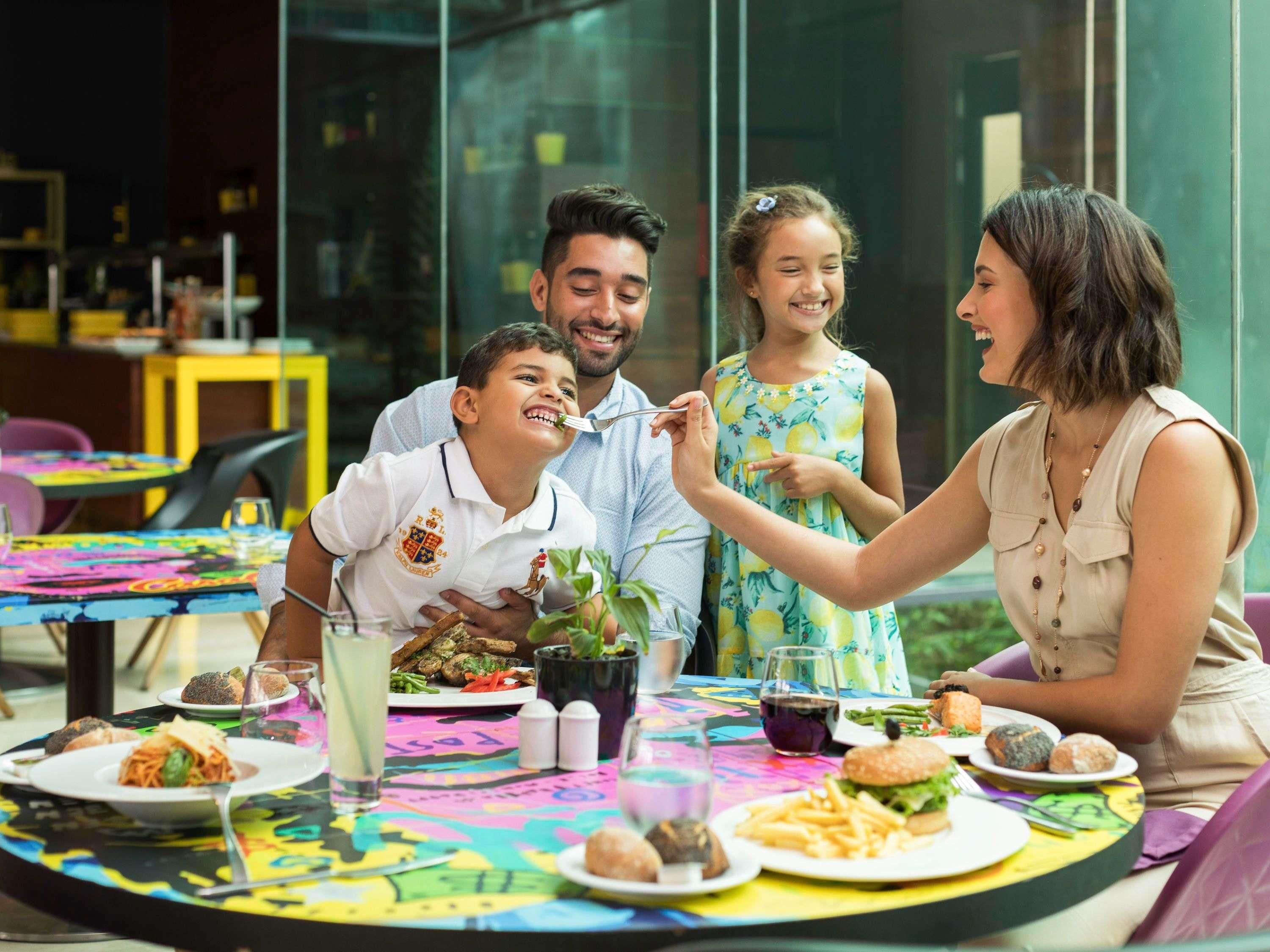 Uma família jantando à mesa com diversos pratos, bebidas e uma toalha colorida.