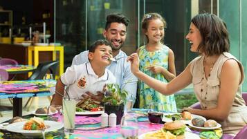 Uma família jantando à mesa com diversos pratos, bebidas e uma toalha colorida.