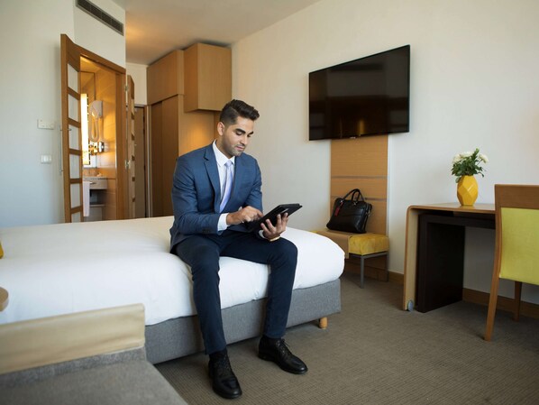 Un hombre trajeado sentado en una cama en una habitación de hotel, con un televisor, una mesita con un florero y una silla.