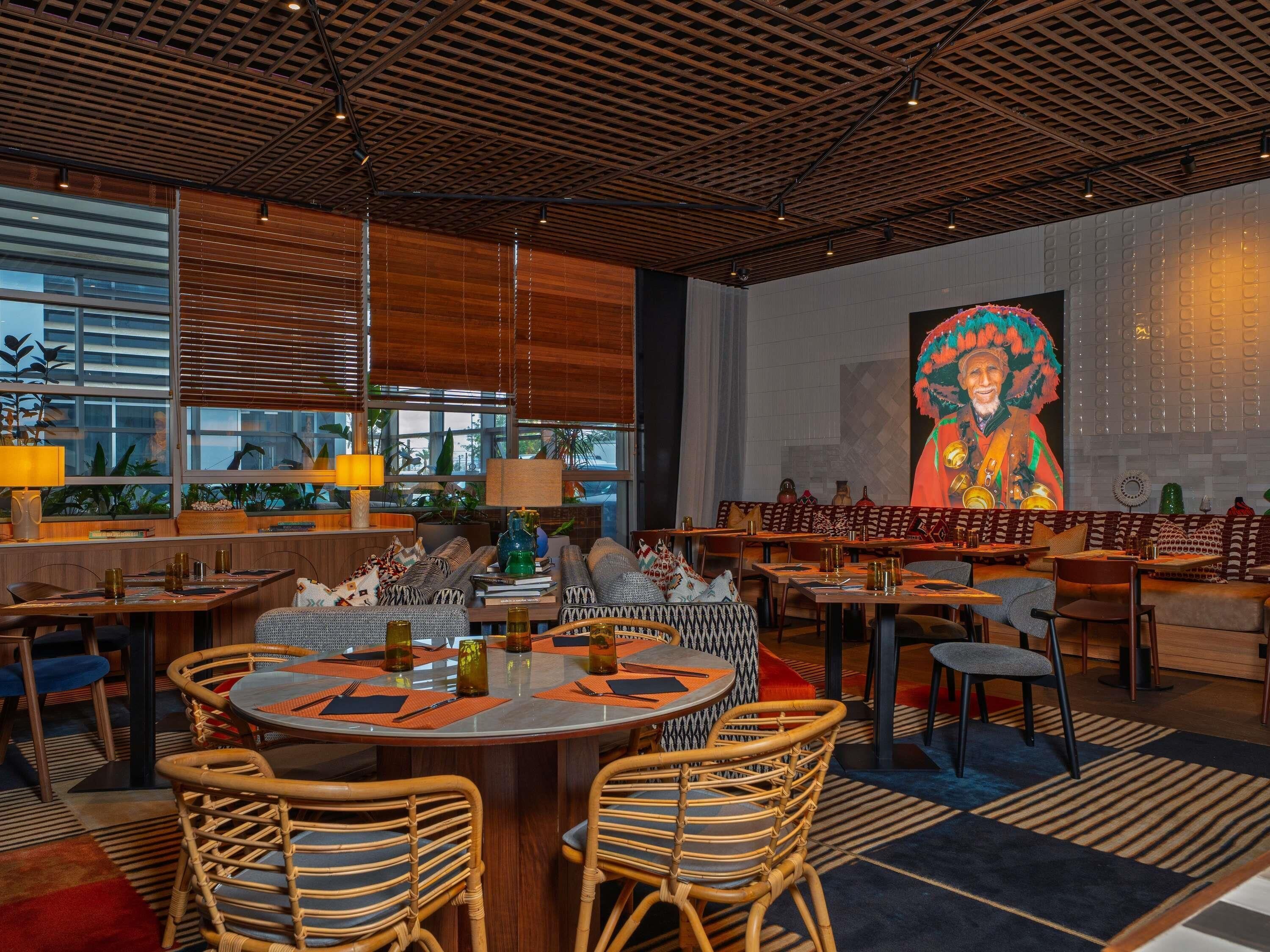 A modern dining area with round tables, wicker chairs, and a large mural on the wall.