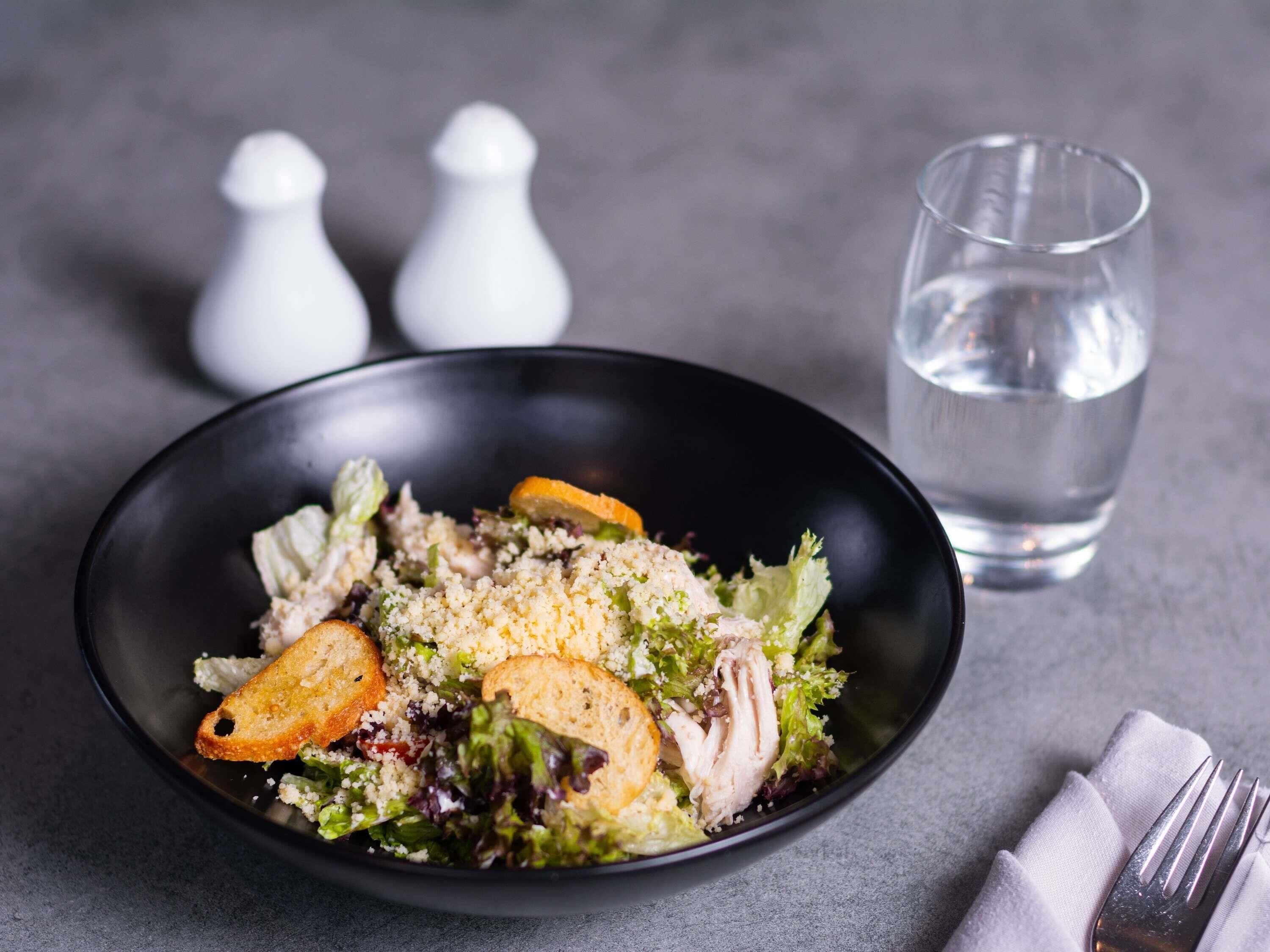 Un saladier noir contenant une salade avec des croûtons et de la laitue, accompagné d’un verre d’eau et de deux salinières (une pour le sel, une pour le poivre) posées sur une surface grise.