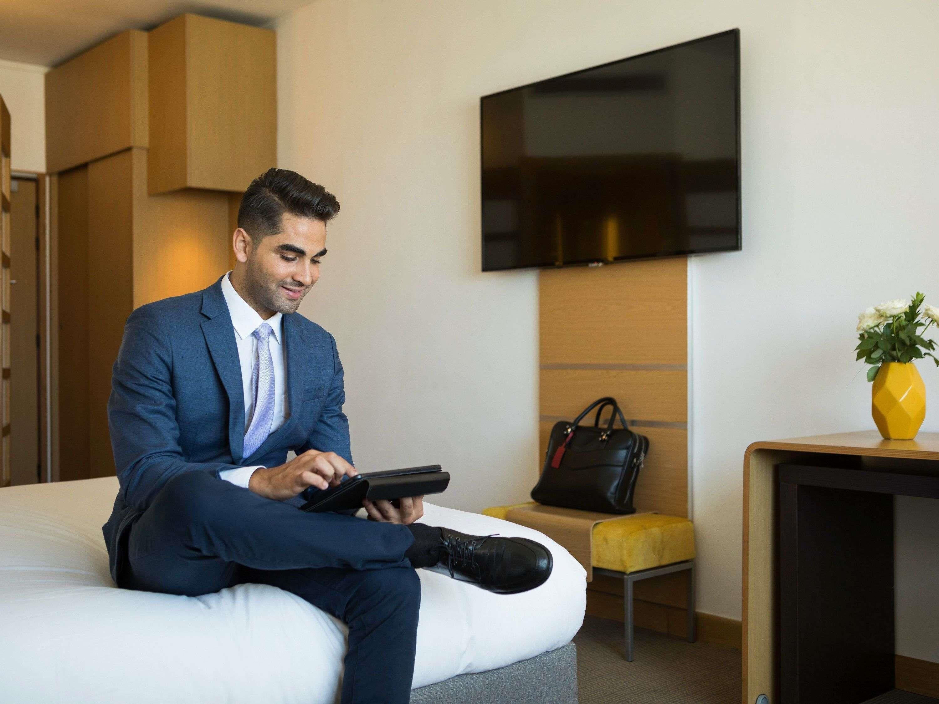 A man in a blue suit sitting on a bed in a hotel room with a TV mounted on the wall, a small table with a vase of flowers, and a chair.