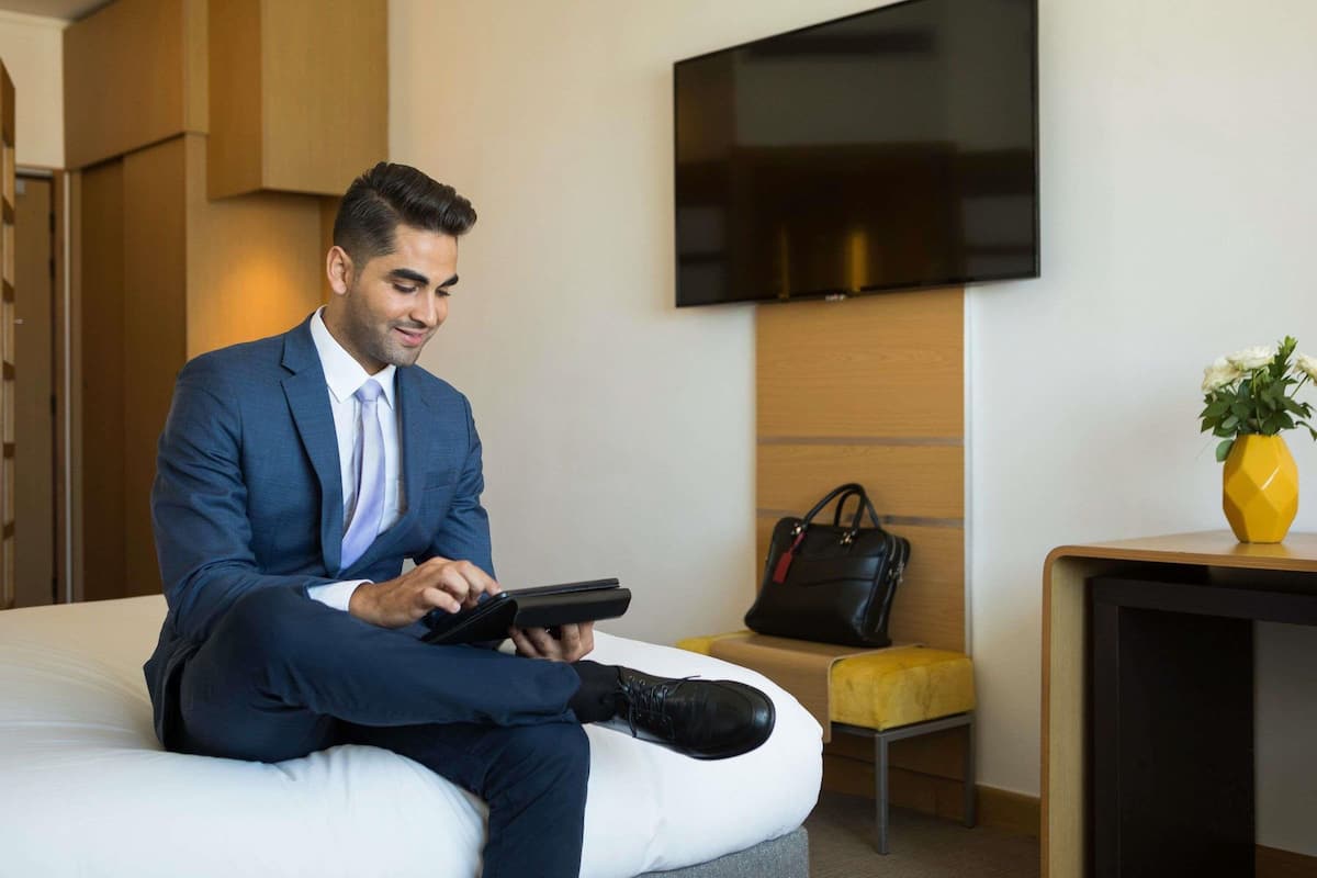 Un homme en costume bleu est assis sur un lit dans une chambre d’hôtel. On peut voir un téléviseur fixé au mur, une petite table avec un vase de fleurs et une chaise.