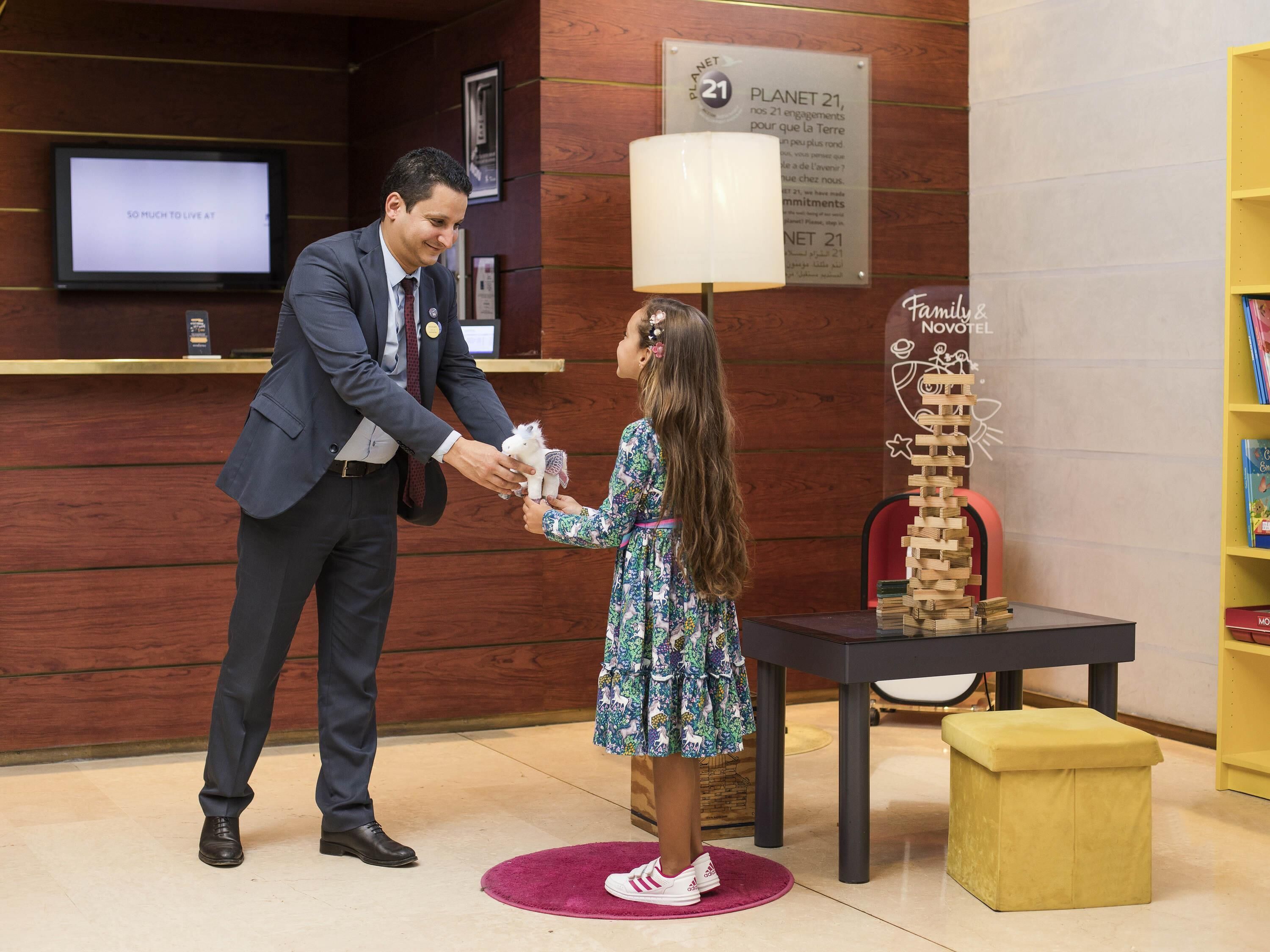 A man handing a stuffed toy to a child in a hotel lobby with wooden paneling, a television, and a Planet 21 sign.